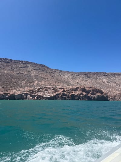 A horizontal outdoor shot depicts a sheer, gray-brown cliff face on the left side and turquoise ocean water on the right. The sky above the cliff is clear blue. The ocean water is disturbed by white froth, suggesting a boat is moving through the water. The location is Unknown, Mexico.