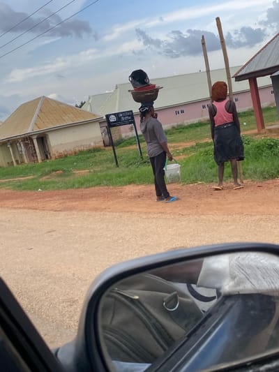 A woman carrying a stack of bowls on her head, dressed in a grey top and black pants, walks on a dirt road in Ban, Nigeria. She holds a white bucket in her left hand. To her right, another woman stands facing away from the camera, wearing a pink tank top and a black skirt, with a wooden pole behind her. In the foreground, a car's side mirror reflects a person wearing a cap. The background shows several buildings with distinct rooflines, a green grassy area, and a cloudy sky. A sign with the text "Delice Bites" is visible to the left of the women.