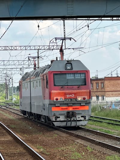 The image shows an electric locomotive on a railway track in Selyatino, Russia. The locomotive is red and gray and has the number "2306-767" visible on its front. It is surrounded by overhead power lines and railway infrastructure. In the background, there are brick buildings and trees. The sky is cloudy. The scene appears to be an operational railway yard or station.