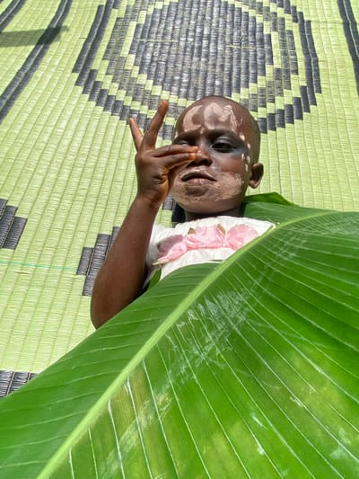 A child with black and white face paint is lying on a light green mat with black geometric patterns. A large, green leaf partially covers the child, revealing a small white and pink floral garment beneath. The child's hand is raised near their face. The location is Ufueseni, Nigeria.