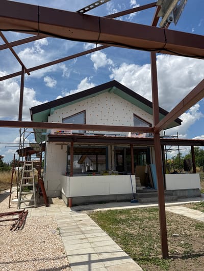A construction site in Graf Ignatievo, Bulgaria, features a house under renovation, with exposed metal beams and scaffolding. The house's exterior walls are covered in white insulation panels, and some windows are visible. A tiled walkway and gravel area are present in the foreground, with some vegetation growing. The sky is blue with scattered clouds, suggesting fair weather. The scene appears to be a daytime shot, likely late morning or early afternoon, given the lighting and shadows. There is no visible text in the image. The overall impression is of ongoing construction work in a semi-rural or suburban setting.