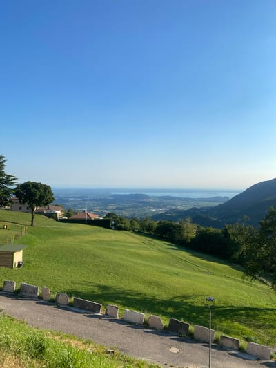 This media file depicts a scenic landscape view from a hilltop. The scene is dominated by a vast expanse of clear blue sky on the left, transitioning to a hazy horizon where a body of water, likely a lake, is visible. Below the sky, a sprawling green meadow slopes downwards, casting long shadows from unseen trees. In the distance, a valley with agricultural fields and scattered buildings is visible.
Along the right side of the frame, a paved pathway runs along the edge of the meadow, bordered by a low stone wall made of irregular rectangular stones. A street lamp with a circular fixture stands on a pole next to the path. Further into the meadow, a cluster of trees is present, with one prominent tree in the foreground. Some low wooden structures with colorful elements are also visible in the lower portion of the image, suggesting a recreational area.
The time of day appears to be late morning or early afternoon, judging by the bright sunlight and the length of the shadows. The weather seems clear and sunny, with a slight haze obscuring the far distance. The location cues, including the mountainous backdrop and the presence of a large lake, combined with the provided location context of Caprino Veronese, Italy, suggest a mountainous region in Northern Italy, possibly overlooking Lake Garda. There are no people or animals clearly visible in the scene. The overall mood is serene and picturesque, highlighting the natural beauty of the location. No visible text is present in the image.