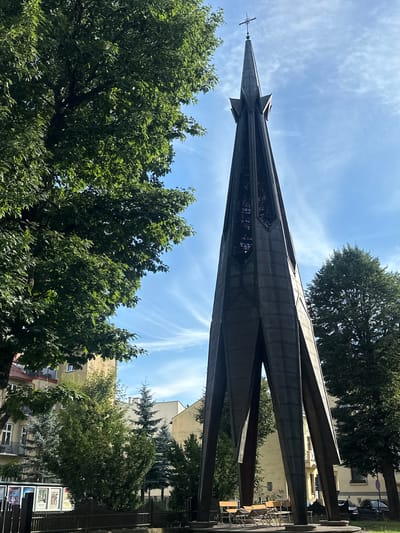 The image captures the Monument to the Fallen and Murdered in the East in Nowy Sącz, Poland. The monument is a tall, dark metal spire with an open, angular design, culminating in a cross at its apex. It is positioned in a public space, with trees on either side and behind it. A large, leafy tree dominates the left side of the frame, its branches extending towards the spire. In the background, several buildings, typical of a European city, are visible, some with apartment windows and balconies. The sky is bright blue with wispy white clouds, suggesting a clear day. In the foreground, at the base of the monument, a circular platform is visible, with several empty chairs and small tables arranged on it. The ground around the monument is covered in grass. The overall scene conveys a sense of solemnity and remembrance.