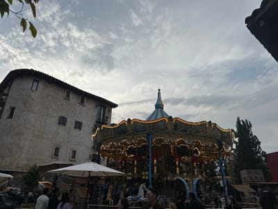 A carousel sits illuminated in front of a building with a tiled roof. The sky overhead is cloudy and overcast. The carousel is crowded with people. An open umbrella stands nearby. To the right, tall green trees provide a backdrop, along with a red building. The building on the left has several small windows.