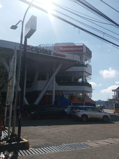 This image depicts a multi-story commercial building, identified by prominent signage as "Cipiaz Cipanas" on the lower facade, "Ramayana" on the upper-middle section, and "XXI" on the top right corner. The building features a modern design with large, angled support columns and multiple levels with balconies. Two blue tents and one multi-colored tent with "GOKANA RAMEN & TEPPAN" signage are set up on the ground level in front of the building.

In the foreground, a black street lamp post with a rectangular sign box stands on the left. Overhead utility lines crisscross the bright, clear blue sky, which exhibits a strong sun flare near the top center. The ground is a light-colored paved surface, likely a parking lot, where a black SUV and a silver SUV are parked. A concrete drain cover with parallel slats is visible at the bottom edge of the image, next to a yellow-painted curb and some scattered litter. To the far right, a small, light-colored building, possibly a gatehouse or ticket booth, is visible. Distant hills and foliage are discernible in the background. No people are visible. This scene is depicted in the city of Ban Phonhxavanh, Laos.