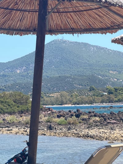 An eye-level, medium shot captures a tranquil beach scene in Lichada, Greece, dominated by the natural beauty of the landscape and the simple comforts of a vacation setting. In the foreground, a wooden pole supports a thatched umbrella, its frayed edges casting dappled shadows. To the left, a partially visible lounge chair with a tan fabric seat is positioned near the water. A scuba diving mask and snorkel lie beside the wooden pole.

The middle ground reveals a rocky shoreline with sparse vegetation, leading to the vibrant blue waters of the sea. Small boats dot the water, and in the distance, a sandy beach is lined with white umbrellas and lounge chairs, indicating a popular tourist spot. Further back, a densely forested mountain rises majestically, its slopes covered in lush greenery. The summit of the mountain is dotted with what appear to be communication towers. The sky above is a clear, bright blue. The overall impression is one of peaceful relaxation and natural splendor.