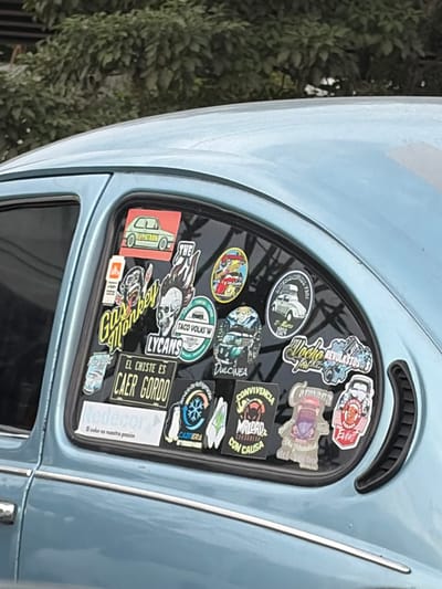 A close-up shows a light blue Volkswagen Beetle's rear window adorned with numerous stickers. Sticker themes include car clubs, car-related humor ("EL CHISTE ES CAER GORDO"), events, and brands ("Redecor"). Specific phrases on the stickers are "TACO VOLKS W," "LYCANS," "DULCINEA," "CONVIVENCIA MALDAD," "WOCHO REVOLKSTOSS." One sticker mentions a "Dia Mundial del cho" event in March 2024. The overall impression is of a car enthusiast who is proud of their vehicle and partakes in VW-related activities.