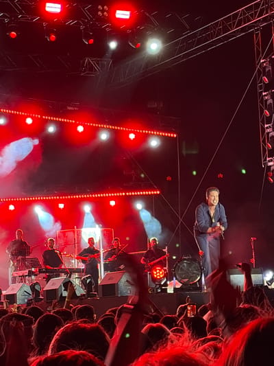 A live music performance is taking place on an outdoor stage at night, lit by numerous bright red lights. The crowd in the foreground is illuminated by the stage lighting, with many people holding up their phones. On stage, several musicians are visible, playing instruments such as keyboards and string instruments. A lead singer, dressed in a dark blue shirt and pants, is in the center of the stage, singing and holding a microphone. The background features a large drum and additional stage equipment. This event is identified as taking place in Alexandroupolis, Greece.