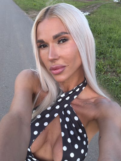 A close-up, diagonally oriented portrait features a young adult female outdoors in what appears to be Конь, Russia. She has long, straight, platinum blonde hair, parted slightly to one side and cascading over her shoulders. Her face is angled towards the camera, and she maintains direct eye contact with a neutral, perhaps confident, expression. She has prominent dark eyebrows and her eyes are enhanced with dark makeup. Her lips are full and painted with a matte pinkish-nude lipstick. She wears a black top or swimsuit adorned with large white polka dots, styled with a halter neckline that reveals her shoulders and décolletage. A script tattoo is visible on her left shoulder/collarbone area, and a smaller, less distinct tattoo can be seen on her right upper arm. The background consists of a gray paved surface in the foreground and green grass with some dirt patches beyond, suggesting a park, path, or natural setting. The lighting indicates it is daytime, possibly bright but diffuse, without harsh shadows.