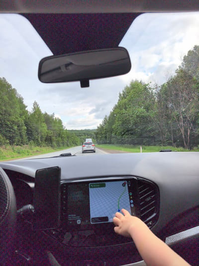 The image captures a first-person perspective from inside a moving car, looking out through the windshield onto a rural road. In the foreground, a child's hand is actively touching the car's central infotainment screen, which displays a navigation map with a highlighted route, a blue pointer, and a "2.2 km" distance indicator. A smartphone in a vertical mount is visible to the left of the screen, near the edge of the steering wheel.
Outside the car, a two-lane road extends into the distance, bordered on both sides by dense, lush green forests under an overcast sky with scattered clouds, indicating a cloudy daytime setting. A silver SUV, equipped with what appears to be a roof box or luggage, is driving ahead in the same lane. Its partially visible license plate, "P005OO", suggests a Russian registration, aligning with the provided location context of Lesnoi, Russia. Another lighter-colored car can be seen further down the road. A white road marker or reflector is visible on the right shoulder of the road. The scene portrays a tranquil moment during a car journey through a wooded landscape.