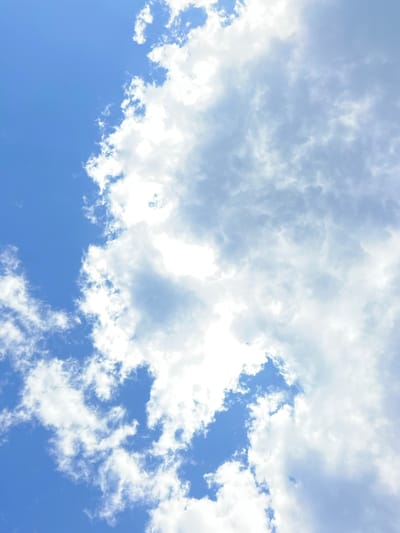 The image shows a close-up view of a bright blue sky with scattered white clouds. The clouds vary in density, with some appearing wispy and translucent, while others are thicker and more opaque, casting subtle shadows. The overall lighting suggests it is daytime, with sunlight filtering through and illuminating the clouds. There is no visible indication of Banská Bystrica, Slovakia, or any specific urban or natural landmarks. The scene is entirely composed of the sky and clouds.