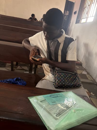 An indoor scene in Osu, Nigeria depicts a young male seated on a dark wooden pew. He has dark skin and short dark hair, wearing a polo shirt that is white on the right side and grey with vertical white stripes on the left, paired with light grey shorts. He holds a gold-colored smartphone with both hands, looking down at its screen. A dark shoulder bag with multi-colored circular and star-like patterns is slung across his body.
In the foreground, on a dark wooden surface, a transparent light green plastic folder contains several papers, including one with a visible blue stamp. To the left of the folder, a crumpled dark blue fabric item is present. In the background, two additional individuals are partially visible seated on similar wooden pews. The room features light-colored walls and a window with vertical bars on the right, admitting natural light. The floor, visible beneath the pews, displays a light-colored patterned or tiled surface.