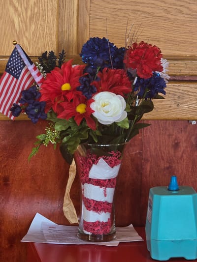 An American flag on a small stick is positioned to the left of a floral arrangement. The floral arrangement consists of red, white, and blue artificial flowers, with a white rose as the centerpiece. The flowers are in a clear glass vase filled with red and white shredded paper. A tan ribbon is tied around the vase. The vase sits on a stack of papers on a red surface. Behind the arrangement, a wooden panel with vertical lines is visible, and to the right, a teal-colored object with a blue nozzle sits on the red surface. The scene appears to be indoors, possibly in a business or event space. The city of Cumming, United States of America is mentioned in the prompt as the location, but there are no visual cues in the image to confirm this specific location.