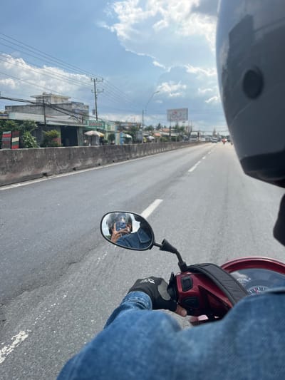A first-person perspective captures a motorcycle ride on a highway. A rider's gloved hand grips the handlebar, and a side mirror reflects the driver taking a selfie with a phone. The scene shows a concrete road, a roadside view of buildings, power lines, and a cloudy sky. The rider wears a helmet, visible at the top of the frame, and a denim jacket. A speedometer is visible.