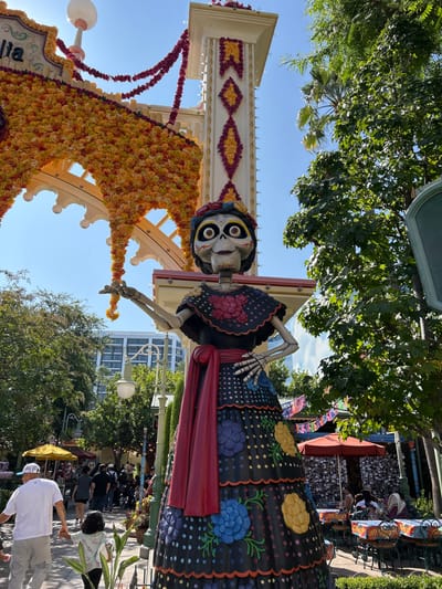 This image captures an outdoor scene within Disney California Adventure Park in Anaheim, United States, specifically depicting the Plaza de la Familia area.

A large, vibrantly colored skeletal female figure, styled as La Catrina, stands prominently in the foreground. The figure has a white skull face with exaggerated eyes and a slight smile, wearing a black blouse with red floral designs, a red waist sash, and a tiered black skirt adorned with white polka dots and large appliqués of blue, yellow, and purple flowers. Its left arm is extended outwards, and its right hand rests on its hip.

Behind the figure, a substantial white architectural archway is heavily decorated with garlands and clusters of orange and red marigold-like flowers, spanning across the upper left portion of the frame under a clear blue sky. Adjacent to the archway, a tall white tower-like structure is visible, featuring red and yellow diamond and banner patterns. Lush green trees flank both sides of the scene, with palm fronds discernible on the right. In the distant background, a multi-story, light-colored modern building with numerous windows is partially visible through the foliage.

On the left side of the image, a paved walkway extends into the mid-ground, populated by several pedestrians. A man in a white t-shirt and light pants walks forward, holding the hand of a small child in a light-colored long-sleeve shirt. To the right, an outdoor dining area is present, featuring red umbrellas, tables draped with patterned tablecloths, and chairs, where multiple individuals are seated. Colorful papel picado banners are strung above this dining area. The entire scene is illuminated by bright daylight.