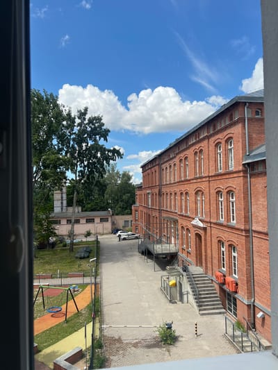 The image depicts an outdoor scene on a sunny day with a bright blue sky and scattered white clouds. On the right side of the frame, a multi-story brick building with numerous arched windows and white frames dominates the view. The building appears to be an older architectural style, possibly a former industrial or institutional structure. To the left of the building, a paved courtyard area is visible, with a playground featuring a swing set and a small slide. Beyond the playground, there is a grassy area with trees, and further back, another smaller building with a light-colored facade and a tall, narrow chimney. Several cars are parked in the courtyard. The scene is located in the city of Oleśnica, Poland, specifically at the former "Wytwórnia Instrumentów Lutniczych" (Luthier Instrument Factory) premises, which is now undergoing redevelopment.