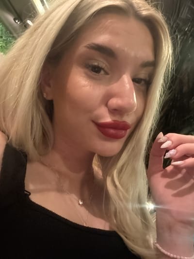 A female individual is depicted in a close-up, chest-up shot. She has long, blonde hair, parted on the left side, with visible roots. Her facial features include dark, well-defined eyebrows, dark eyes, and a prominent nose. Her lips are coated with a dark red or berry-colored lipstick with a glossy finish. A small, clear stud earring is visible in her left earlobe. She is wearing a black top or tank top. Around her neck are two layered necklaces: one delicate chain with a single, clear, circular pendant, and another shorter chain, possibly a paperclip style. Her left hand is raised, with fingers slightly curled, showcasing long, pointed nails. The nails feature a blend of light pink/nude and white polish, with the ring finger adorned with a small, sparkly, snowflake-like embellishment. A bracelet composed of small, light pink beads is visible on her left wrist. The background is dark and out of focus, indicating an indoor or nighttime outdoor setting. Faint green foliage is visible on the far left, and multiple blurred light sources (bokeh) are present in the background, along with a distinct light flare near her left hand. The specific location in the city of Strumica, North Macedonia, is not discernible from the visual information within the image.