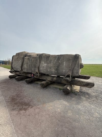 An outdoor scene in Amesbury, United Kingdom, depicts a large, rectangular sarsen stone replica resting on a wooden roller system, simulating ancient transportation methods. The stone, roughly 4-5 meters long and grey with a coarse, textured surface, is secured by four loops of thick, dark braided rope. These ropes encircle the stone vertically, with two loops visible on the front face. The stone is supported by two primary longitudinal dark wooden beams, which in turn rest on six shorter, perpendicular wooden rollers laid directly on a gravel path. A small circular red and white "No climbing" sign is affixed to one of the front wooden rollers. To the left of the gravel path, faint grey structures and sparse trees are visible in the distant background. To the right, an expansive, flat green grass field extends to the horizon. The sky is overcast and uniformly light grey. No individuals are present in the image.