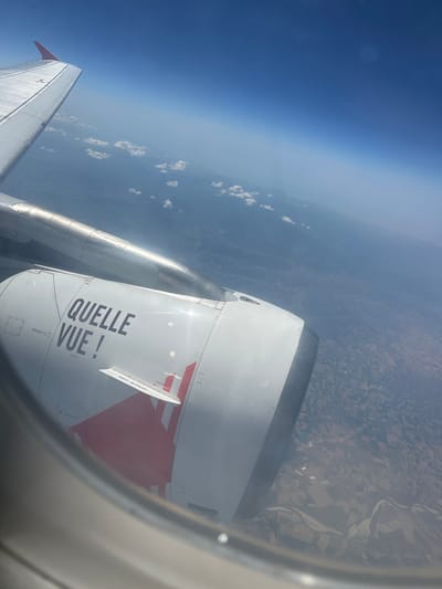 The image is taken from an airplane window, showing a view of the Earth's surface from a high altitude. The scene is dominated by the wing and engine of the airplane, which are white with red accents and some visible text. The sky is a gradient of deep blue to a hazy lighter blue, with some scattered white clouds visible. Below the aircraft, the landscape is a patchwork of agricultural fields, with a meandering river visible in the distance. The time of day appears to be daytime, judging by the bright sunlight. The weather seems clear with some haze at the lower altitudes. The text visible on the engine reads "QUELLE VUE !" which translates to "What a view!" in French, and also "NO STEP". The image appears to be taken over or near Pavia, Italy, given the geographical context provided. The overall impression is one of a scenic aerial view, likely experienced during a flight.