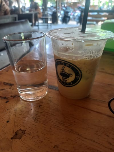 In Gampongaceh, Indonesia, a close-up, eye-level shot focuses on two drinks on a wooden table. To the left, a clear glass contains a light-colored liquid, likely water. To the right, a clear plastic cup with a lid holds an iced coffee with a visible black straw. The plastic cup exhibits condensation on its exterior, indicating a cold beverage. A black circular logo with "KOPI ROASTE" and a stylized coffee cup is emblazoned on the side of the cup. The wooden table surface shows a textured grain and some minor imperfections. In the blurred background, an outdoor seating area is visible with the faint shapes of people and furniture, suggesting a cafe or restaurant setting. The overall lighting is natural and somewhat diffused, hinting at daylight hours.