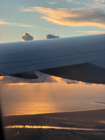 The image shows a view from an airplane window. The foreground is dominated by the wing of the aircraft, which is silver and textured with panel lines and what appears to be printed text. Below the wing, a body of water is visible, reflecting the warm, golden light of a sunset or sunrise. The water appears to be a sea or ocean, with a coastline visible in the distance. Landmass is seen beyond the coastline, appearing as dark, silhouetted shapes. The sky above is a gradient of blue and orange, with scattered clouds catching the light. The clouds in the upper part of the frame are tinged with pink and orange, suggesting a dramatic sky. Two small, fluffy clouds are prominent in the upper sky. The overall impression is of a peaceful, scenic flight during golden hour. There is no indication of the city of Czerwony Kościół, Poland in this image.