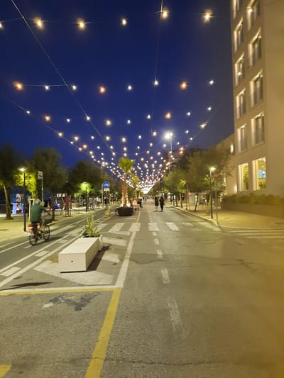 The image depicts a pedestrianized street at dusk, likely in Pesaro, Italy, illuminated by a string of festoon lights overhead. A modern, light-colored building with multiple windows dominates the right side of the frame. The street itself is paved and marked with white lines indicating a pedestrian crossing and possibly a bicycle lane.

In the foreground, a man wearing a green shirt is cycling on the street. To his left, a long, white, rectangular planter with some greenery is positioned. Further down the street, several people are walking, some in groups. Trees line the left side of the street, and a palm tree is visible in a planter on the right. The sky is a deep blue, indicating the transition from day to night. The overall atmosphere is one of a pleasant evening in a public space.