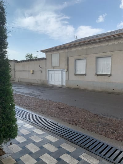 An outdoor, eye-level shot captures a street in Urtaaul, Uzbekistan, with a large building on the right and a concrete wall on the left. A green tree is on the far left. The building, constructed of grey concrete, features a dark, corrugated roof with decorative trim along the facade. Two windows on the building are covered with white sheeting. A large, white double door is visible on the right side of the building, and a smaller, single door is to its left. The street in front of the building is paved with asphalt and shows signs of wear, with patches and puddles, suggesting recent rain. A drainage grate runs along the edge of the street, adjacent to a patterned walkway made of grey and white tiles. The background shows a cloudy, pale blue sky with some faint electrical wires strung across it.