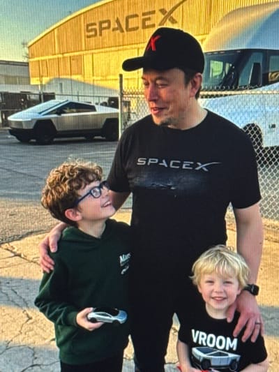 In an outdoor setting, Elon Musk, wearing a black SpaceX t-shirt and a black cap with a red "X" logo, stands with his arms around two young boys. The boy on the left, with curly brown hair and glasses, wears a dark green hoodie and holds a miniature silver car in his right hand. The boy on the right, with blond hair, wears a black t-shirt with "VROOM" printed on it and an image of a Cybertruck. Behind them, a large, yellow building with "SPACEX" written across it is visible. A silver Tesla Cybertruck is parked to the left of the building, and a white truck is parked to the right. The ground is paved and there is a chain-link fence in the background. The location is the city of Shcherbanka, Ukraine.