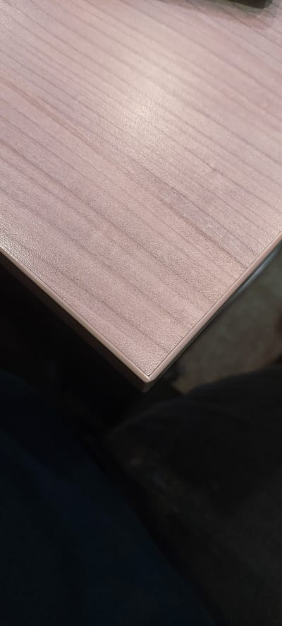 The image shows a close-up view of a table surface with a wood grain pattern. The table has a light pinkish-brown hue and a slightly textured finish. The edge of the table is visible, with a thin, lighter-colored trim. Below the table, a dark, out-of-focus area suggests a person's clothing, possibly dark blue fabric. The lighting highlights the texture and grain of the wood, with some areas appearing brighter due to reflections. The scene appears to be indoors. The location in Pagedangan, Indonesia, is not discernible from the visual information in the image.
