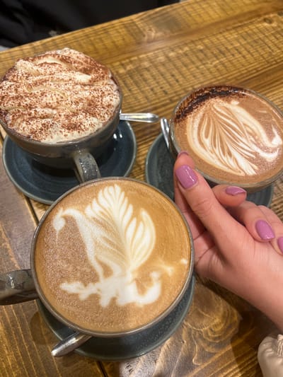 The scene is an overhead shot of a wooden table in an indoor setting, featuring three mugs of hot beverages. A person's hand with light purple nail polish is visible near one of the mugs, with their index finger slightly pointing to it. The mugs are arranged in a triangular pattern, with one containing a drink topped with cocoa powder and foamed cream, another containing latte art, and the third showing latte art in detail. Each mug sits on a dark-colored saucer. The specific location within Mole Valley, United Kingdom, is not discernible from the provided image.
