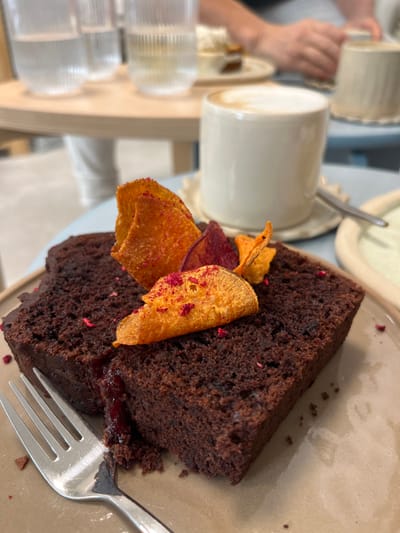 The image displays a dessert setting in an indoor cafe environment. The primary subject is a rectangular slice of dark brown cake, likely chocolate, served on a light brown, rectangular plate. A red-purple fruit filling or jam is visible within a cut section of the cake on the left side. The cake is garnished with several thin, crispy chips of varying orange and reddish-purple hues, which are flecked with small red particles. A silver-colored metal fork is positioned on the left side of the plate, partially inserted into the cake. To the right, a cream-colored, cylindrical mug containing a beverage with a foamy top, consistent with a latte, rests on a light-colored, scalloped-edge saucer. A spoon is partially visible on the saucer. In the blurred background, two clear, fluted glasses filled with water are visible on a light wooden table surface. A person's arm and hand are present in the upper right background, interacting with another object on a separate table. The specific location within Kaiserslautern, Germany, is not discernible from the image content.