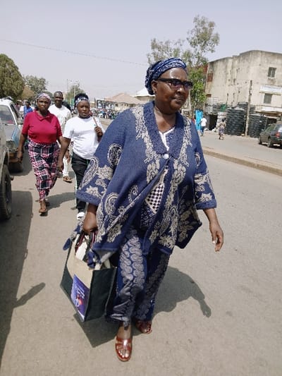 The image captures a daytime street scene in Mumbwa, Zambia. In the foreground, an adult female with dark skin walks forward and slightly to the viewer's right. She is attired in a dark blue patterned tunic top with a v-neck and matching trousers, a blue patterned headwrap, and dark-framed eyeglasses. Her left hand holds a two-tone fabric tote bag, dark green/black and light brown/khaki, which displays a colorful graphic on its side. She wears dark reddish-brown open-toed sandals.
Behind her, positioned to the left and slightly further back, a group of three other individuals follows. The lead individual in this group is a female wearing a red short-sleeved top and a patterned skirt. Following her is another female in a white short-sleeved t-shirt and dark trousers, and behind her, a male in a light-colored, possibly white, long-sleeved shirt and trousers.
The scene takes place on a paved road surface under bright daylight, with clear shadows cast on the ground. To the far left, the front portion of a light-colored vehicle is visible. The background features various buildings lining the street, some appearing multi-story and exhibiting signs of wear. Notably, on the right side of the street, a large, light-colored building stands, with multiple stacks of large black plastic tanks positioned outside it. Sparse trees and power lines are also visible in the distance. Other pedestrians are faintly discernible further down the street.