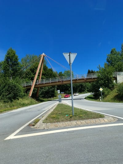 The image captures a clear, sunny day at an intersection featuring a pedestrian bridge. The bridge, constructed with a modern design incorporating orange cables and a light-colored deck, spans over a two-lane asphalt road. Two cars are visible on the road: a red car in the left lane and a white car in the right lane, both moving away from the viewer. The intersection is marked with white road lines, and a triangular yield sign is prominently placed on a metal pole in the foreground. Green trees and vegetation line the sides of the road and surround the bridge. A yellow diamond-shaped sign is visible on a pole to the right of the intersection. The scene suggests a rural or semi-rural setting, likely within or on the outskirts of Freyung, Germany, given the information provided.