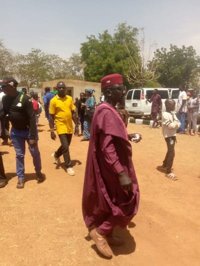 This image depicts an outdoor gathering of individuals on a sunny day in Kagoma, Nigeria. The ground is reddish-brown dirt. In the foreground, an older male with dark skin, wearing a maroon traditional long-sleeved tunic over matching trousers and a maroon cap, walks towards the left of the frame. He has sunglasses, a dark beard, and holds a crumpled white item in his right hand. He wears brown loafers. To his left, slightly behind, a man with dark skin wearing a yellow short-sleeved shirt, dark trousers, and light-colored sneakers walks forward. He has beaded bracelets on his left wrist. Further left, a man in a black long-sleeved shirt, blue jeans, and a dark baseball cap with green and yellow accents is partially visible, walking left.
In the midground, a diverse group of people, including men, women in patterned dresses, and children, are dispersed. A young boy in a light-colored long-sleeved shirt, dark patterned trousers, and sandals stands with his arms crossed to the right of the maroon-clad man. A white passenger van is parked behind this group, facing left. Behind the van and people, a light-colored, single-story building with a flat roof is visible. The background features several green trees with varying foliage density against a clear, light blue sky. One tall, bare-branched tree stands prominently among the green foliage. The scene is brightly illuminated by natural sunlight, suggesting midday.
