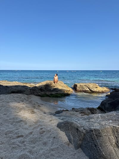 A full shot captures a woman in a pink bikini sitting on a rock formation on a beach. She is looking to her right, with her knees bent and arms resting on them. The beach has sand and more rock formations. The sea is blue and calm, with yellow buoys scattered in the distance. The sky is clear and blue. The location is Platja de Sant Pol, in the municipality of Sant Feliu de Guíxols, on the Costa Brava, within the Baix Empordà region, in Catalonia, Spain.