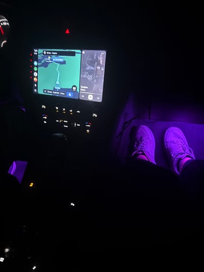 The image depicts the interior of a vehicle at night, specifically from the perspective of a passenger in Zdravets, Bulgaria. The scene is dominated by low light, augmented by purple ambient lighting in the footwells and illumination from the vehicle's infotainment system and controls.

In the center of the dashboard, a wide infotainment screen is visible. The left side displays a navigation map indicating a route with an estimated distance of 12 km and a travel time of 12 minutes to a destination marked "Асен," with a speed limit of 68 km/h. Text "Мин Кубрат" is also partially visible on the map. The right side of the screen shows a music player interface, displaying album art with a person, the title "Трафик" (Traffic), and the artist "Selina." A play/pause icon is visible below. Above the screen, a red hazard light button is illuminated.

Below the infotainment screen, the vehicle's climate control panel is illuminated, showing temperature settings of "24°" on both the left and right sides. Buttons for AUTO, SYNC, and AC are also visible. A gear shifter is partially discernible further down the center console.

In the foreground, extending from the lower right into the center, are the feet and lower legs of a person, presumably the passenger. The person is wearing light-colored, laced sneakers, which appear purple due to the ambient lighting in the footwell. The feet are resting on the floor, indicating a relaxed posture. The lower legs are clad in dark trousers or clothing. The overall impression is that of being inside a modern vehicle during nocturnal travel or a stop.