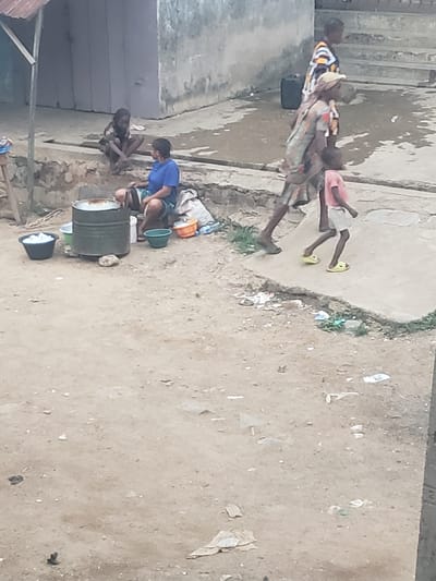 The scene depicts an outdoor area in Alalubosa, Nigeria. Several children are present. Some are washing clothes using buckets and a metal barrel, while others are standing or walking nearby. The ground is sandy and littered with debris. There are buildings and a set of stairs in the background.