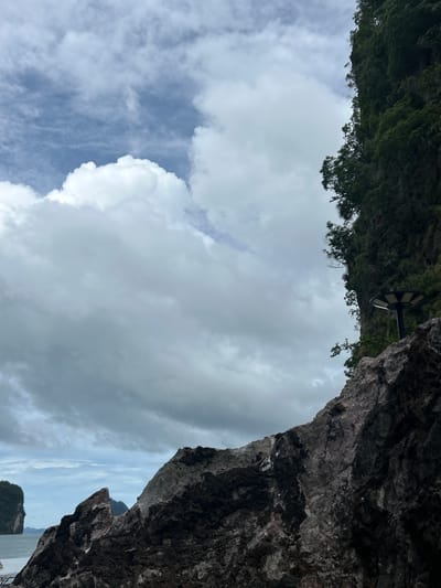 The image depicts a rocky coastline with lush vegetation on a steep cliff face under a cloudy sky. In the foreground, dark, rugged rocks dominate the lower portion of the frame, their textured surfaces detailed. To the right, the dense green foliage of the cliff obscures much of the rock. A modern, dark-colored solar-powered lamp is perched on the edge of the rock, overlooking the water. In the background, islands are visible in the distance, their forms softened by the hazy atmosphere and the overcast sky, which is filled with large, billowy white clouds interspersed with patches of blue. The scene conveys a sense of natural grandeur and remoteness.