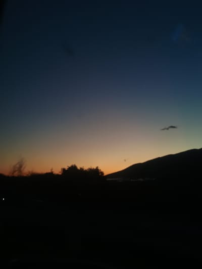 The image captures a dusk scene in Vetren, Bulgaria. A darkened landscape with trees is silhouetted against a gradient sky transitioning from dark blue to orange. The upper part of the image shows a silhouette of mountain.