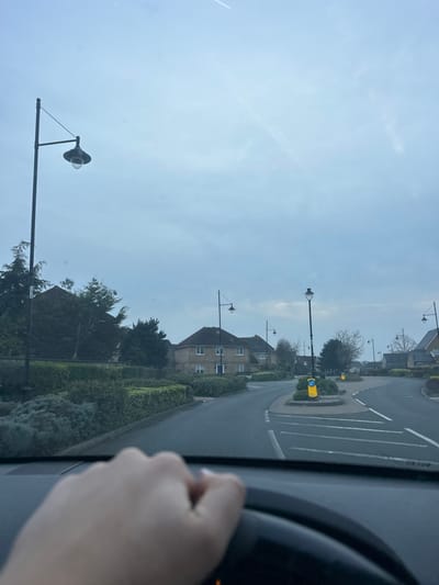 The image depicts a residential street in Gillingham, United Kingdom, captured from the interior perspective of a vehicle. A left hand, featuring light-colored fingernail polish on the thumb and index finger, grips a black steering wheel in the lower-left foreground. The vehicle's dashboard is partially visible below the windshield.
The scene ahead shows an asphalt road curving slightly to the left, leading into a multi-pronged junction or small roundabout. The central island of this junction contains low, manicured hedges and two yellow cylindrical bollards with reflective white bands. White road markings are present on the asphalt.
Several two-story detached or semi-detached houses with light-colored brick facades and dark tiled roofs are visible in the midground and background, set amidst green hedges, bushes, and trees. Multiple black streetlights with fluted, traditional lantern-style tops are spaced along the roadside. The sky is overcast and light blue-grey, occupying the upper half of the frame. A faint white aircraft contrail is discernible in the upper right quadrant of the sky. No other individuals or active subjects are present. The environment suggests a modern, well-maintained suburban area.
