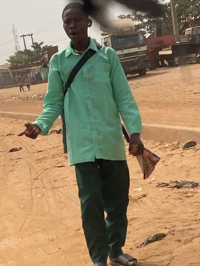 An outdoor scene in Lapai, Nigeria, depicts a young dark-skinned male standing on an unpaved, dusty, light brown road. He wears a light green long-sleeved collared shirt, dark green trousers, and dark grey slides. A black strap, likely from a bag, crosses his chest diagonally from his right shoulder. A white earbud is visible in his right ear. His mouth is slightly open. His left arm is extended forward and slightly to his left, with the index finger pointed. His right hand clutches crumpled fabric or paper, and he wears a ring on one finger of that hand. The ground exhibits tire tracks and scattered debris. In the mid-ground, a white and red DAF heavy truck is visible, partially obscured, along with other industrial machinery components. In the far background to the left, several indistinct figures are standing near low structures, with utility poles and power lines also visible. The environment appears arid under bright daylight conditions.