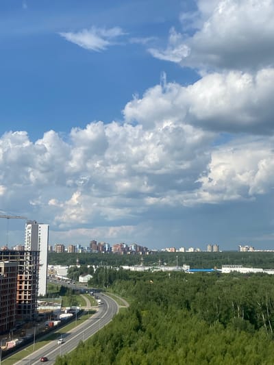 An aerial, high-angle, full outdoor shot captures a cityscape of Kazan, Russia, under a partly cloudy sky. A large, partially constructed building with exposed rebar and concrete dominates the left side of the frame, accompanied by a construction crane. Adjacent to it stands a finished, multi-story building with a white facade. A highway with several vehicles, including cars and trucks, curves through the scene, bordered by a dense forest of green trees. In the distance, a cluster of modern residential buildings forms the city skyline, nestled amongst more greenery. The sky is a vibrant blue with scattered white and grey clouds, some of which appear dark and heavy.