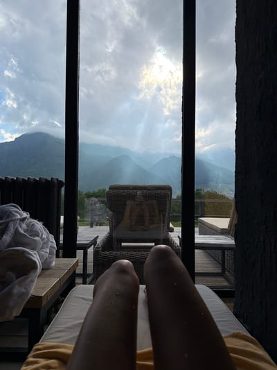 The image shows a person's legs resting on a lounge chair, with a view of mountains and a cloudy sky through a window. The scene is set outdoors, likely on a balcony or patio, with outdoor furniture visible. A white towel is draped over the side of the lounge chair. The lighting suggests it might be daytime, possibly early morning or late afternoon, given the soft, diffused light. The weather appears overcast with patches of sunlight breaking through the clouds. The mountainous landscape suggests a location in a rural or remote area, consistent with the provided location context of Stepantsminda, Georgia. The overall mood conveyed is one of relaxation and enjoyment of a scenic view. There is no visible text in the image.