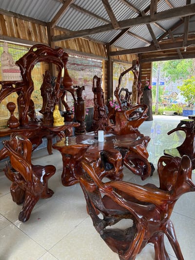 The image depicts an outdoor display area in Hòa Đa, Vietnam, featuring elaborately carved wooden furniture and decorative statues. The furniture, primarily tables and chairs, are crafted from gnarled, polished wood, showcasing organic shapes and rich, glossy finishes. Several statues, including a seated Buddha figure and other human-like carvings, are displayed on and around the furniture. Pink flowers are visible in vases and arrangements. The scene is partially covered by a corrugated metal roof supported by wooden beams and planks, suggesting an open-air pavilion or market stall. The flooring is tiled with a light-colored checkered pattern. In the background, greenery and trees are visible, indicating an outdoor environment. The overall impression is that of an artisan's display or a shop selling unique wooden art pieces.