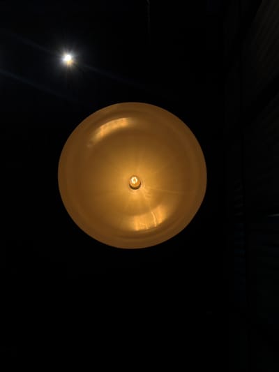 The image, captured in Fraccionamiento Los Heroes Queretaro, Mexico, depicts an overhead perspective dominated by artificial light. A large, circular, convex light fixture, golden-yellow in color, occupies the central and lower portions of the frame. An exposed, glowing filament bulb is visible at its precise center, casting warm, amber light and creating reflections on the fixture's surface. In the upper-left quadrant, a smaller, intensely bright white light source is present, emitting distinct lens flares. The background is predominantly dark. Faint, dark, vertical lines or slats are discernible in the upper-right corner, suggesting a textured wall or blinds. A thin, dark line, possibly a chain, is barely visible descending from the top edge, positioned above the main light fixture. No individuals or explicit actions are present.