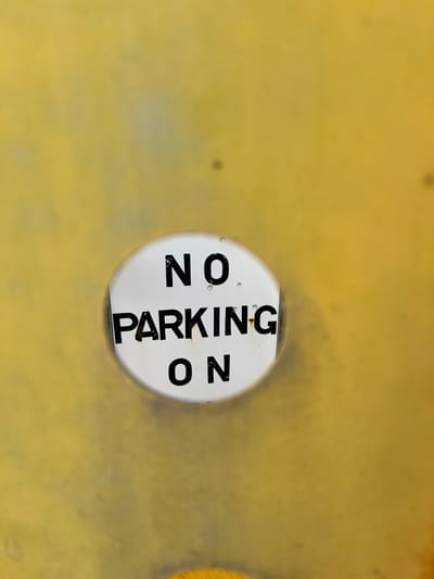 This is a close-up, eye-level, outdoor, medium shot of a white circular sign that reads "NO PARKING ON" in black text. The sign is affixed to a textured yellow surface, possibly a wall or a metal barrier. The yellow surface appears weathered with some faint smudges and variations in shade. The sign itself has a few small water droplets or specks on its surface. The lighting suggests it is daytime. The image does not provide any information about the city of Terhillion, Ireland, or any people or actions.
