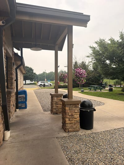 An outdoor scene features a building with stone and light-colored siding, under a covered porch with wooden beams and a single light fixture. A security camera is visible on the side of the building. To the left of the building, a blue newspaper dispenser stands on a concrete walkway. The walkway leads to a seating area with picnic tables and trash cans. In the background, a white SUV is parked near a sign that reads "AIR." Trees and foliage surround the area, and the sky is overcast. The image appears to be taken in Union, United States of America.
