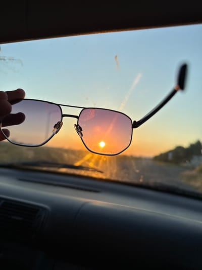 The image depicts a close-up of a pair of sunglasses held by a hand, with the setting sun reflected in the lenses. The sunglasses are aviator-style with dark frames and tinted lenses that show a gradient of pink and purple hues. The sun, a bright orb, is prominently visible in the upper lens, casting a warm glow and creating lens flare effects. The hand holding the sunglasses is partially visible at the bottom, with fingers gripping the lower frame.

The scene appears to be set inside a vehicle, suggested by the interior elements visible on the right side of the image, which include a dark dashboard and possibly a car window. The background, seen through the sunglasses and the car window, shows a blurred landscape with a road and some foliage under a clear, pale blue and orange sky, indicative of either sunrise or sunset. The overall lighting and the colors in the sky and reflections suggest that it is either dawn or dusk.

There is no visible text in the image. The mood conveyed is one of peaceful contemplation or enjoying a scenic moment during travel, possibly reflecting a sense of calm or appreciation for the natural beauty of the sunset. The location context provided is Tankovo, Bulgaria.