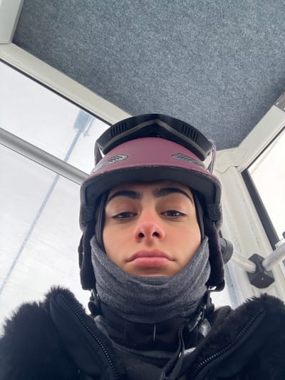 The image presents a close-up, head-and-shoulders view of an individual inside an enclosed cabin, consistent with a ski gondola or cable car, situated in the ski resort area of Valtournenche, Italy. The subject, a young adult with dark hair, prominent eyebrows, and dark eyes, faces the camera with a neutral expression; their nose shows a slight reddish tint. They are equipped for cold weather, wearing a dark reddish-maroon ski helmet with black goggles resting on top. A dark grey neck gaiter covers their lower face and neck, and a black winter jacket with a visible fuzzy or fur-lined collar is worn over a dark inner garment. The cabin's interior features a light grey, textured ceiling and white structural framing. Through a large window to the subject's left, a blurry, light grey exterior indicates snowy or overcast conditions, with faint horizontal lines suggesting distant ski lift elements. A silver horizontal bar is mounted to the cabin's interior wall on the right.