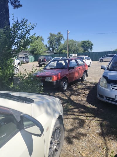 The image depicts an outdoor parking area in Shimanovsk, Russia. Several vehicles are visible, including a red hatchback in the foreground, a silver sedan to its right, and other cars and SUVs parked further back. The ground is covered with gravel and pine needles, and trees and bushes provide shade. A light pole stands in the background. The sky is clear and blue. The scene appears to be a typical day in a parking lot, possibly near a residential or commercial area.